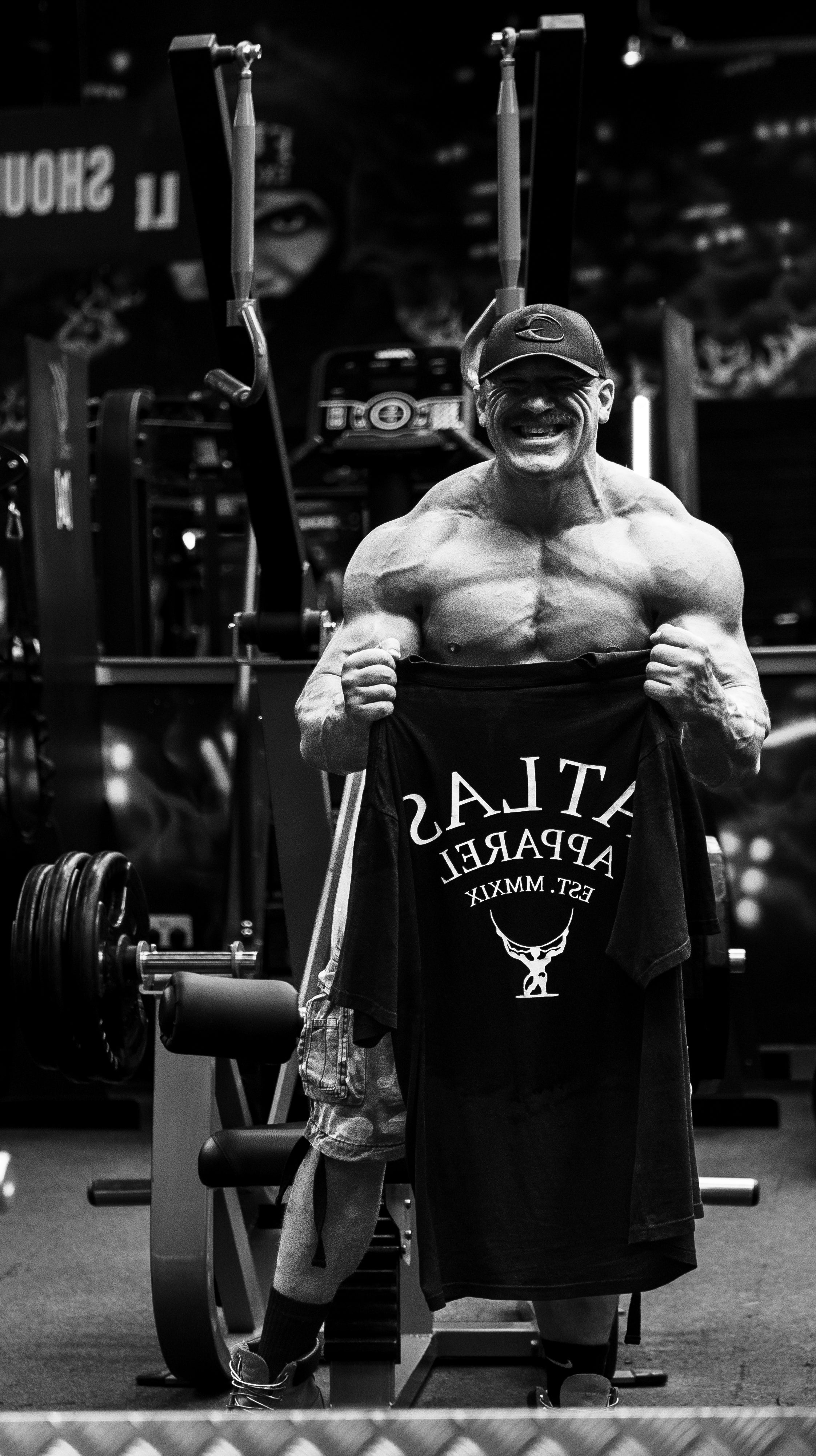 Shirtless man holding up the black oversized t shirt in front of a gym mirror.